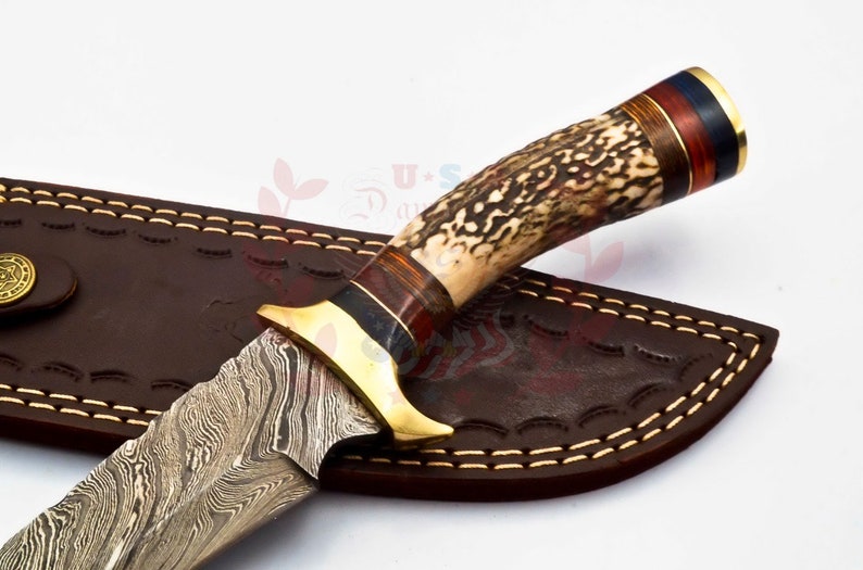 Puede incluir: Un cuchillo de caza con una hoja de acero damasco y un mango de asta de ciervo marr&oacute;n con detalles dorados. El cuchillo est&aacute; en una funda de cuero marr&oacute;n con costuras.