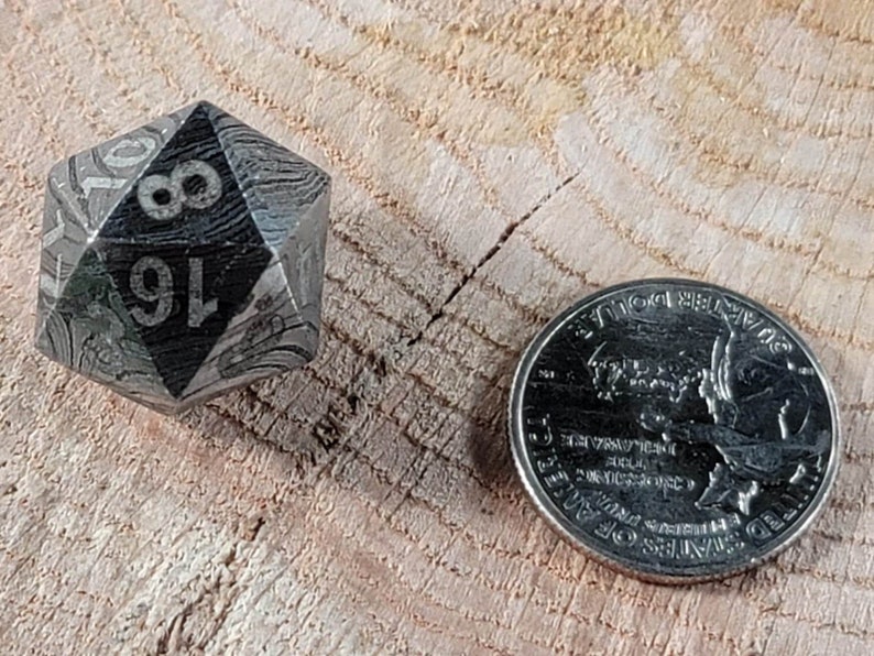 May include: A silver metal d20 dice with black numbers and a swirling pattern. The dice is sitting on a wooden surface next to a US quarter.