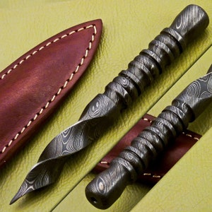 May include: Two twisted, silver-colored, metal daggers with a patterned finish. The daggers are lying on a green leather surface. One dagger is partially covered by a brown leather sheath.