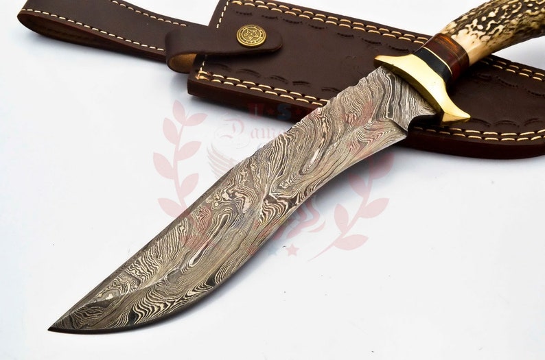 Puede incluir: Un cuchillo de caza con una hoja de acero Damasco y una funda de cuero marr&oacute;n. El mango est&aacute; hecho de asta de ciervo y tiene un acento de lat&oacute;n.