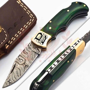May include: A folding knife with a green handle and a patterned blade. The knife features a gold-colored bolster with the text "Bodies Graves". A brown leather sheath is visible in the background.