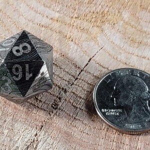 May include: A silver and black metal d20 dice with a textured surface and the numbers 1 through 20 etched on the faces. The dice is about the size of a UK 20 pence coin.