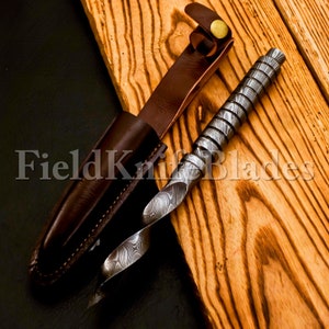 Könnte beinhalten: Ein gedrehtes, damaszener Stahlmesser mit einer braunen Lederscheide. Das Messer hat ein einzigartiges, spiralförmiges Design und ist scharf. Die Scheide hat eine Gürtelschlaufe und einen Druckknopfverschluss.