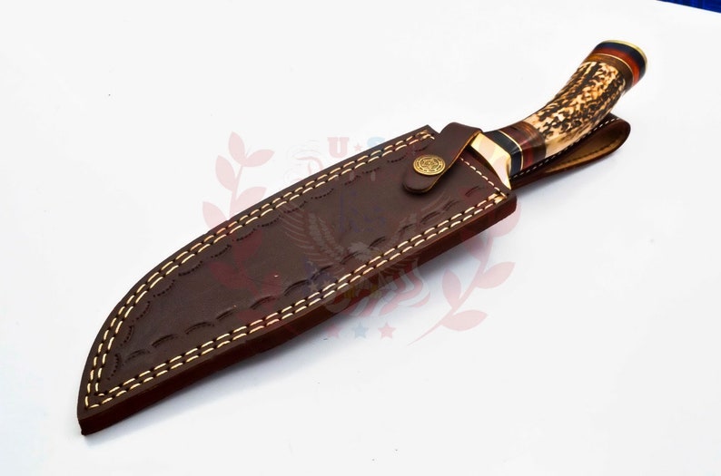 Puede incluir: Una funda de cuero marr&oacute;n con costuras decorativas y un bot&oacute;n de lat&oacute;n. Un cuchillo de caza con mango de asta de ciervo marr&oacute;n y blanco est&aacute; dentro de la funda.