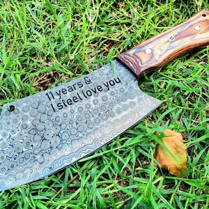 May include: A large, engraved,  damascus steel cleaver with a wooden handle. The blade has a hammered finish and the handle is made of wood with a dark brown stain. The blade is engraved with the words "11 years & I steel love you".
