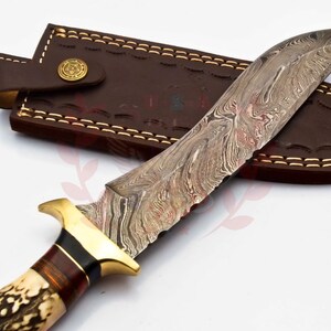 Puede incluir: Un cuchillo de caza con una funda de cuero marr&oacute;n. El cuchillo tiene una hoja con patr&oacute;n y un mango de asta de ciervo marr&oacute;n y blanco.