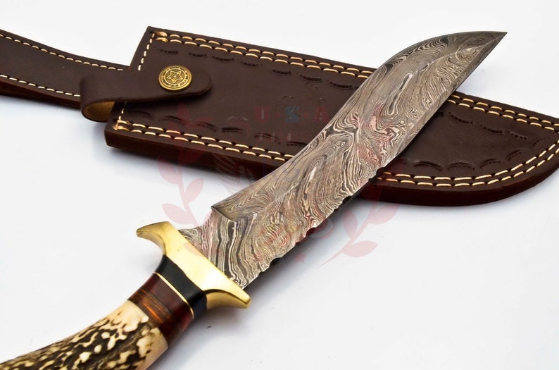 Puede incluir: Un cuchillo de caza con una funda de cuero marr&oacute;n. El cuchillo tiene una hoja con patr&oacute;n y un mango de asta de ciervo marr&oacute;n y blanco.