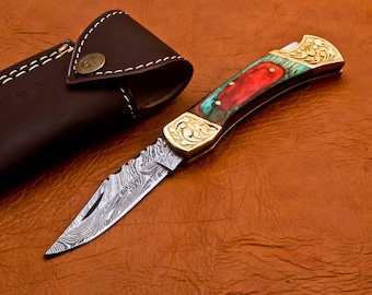 Puede incluir: Un cuchillo plegable de bolsillo con una hoja de acero Damasco y un mango de madera de colores. El mango está decorado con detalles dorados y motivos florales. El cuchillo viene con una funda de cuero marrón.