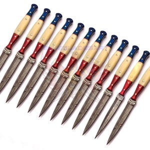 May include: A collection of eleven decorative knives with a patriotic color scheme. The handles are cream-colored with red and blue accents, and the blades have a patterned design. The knives are arranged on a white surface.