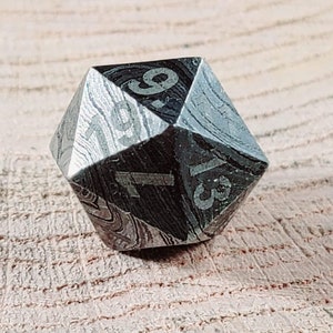 May include: A black and silver metal d20 dice with numbers 1 through 20 etched on each side. The dice has a textured, marbled pattern.