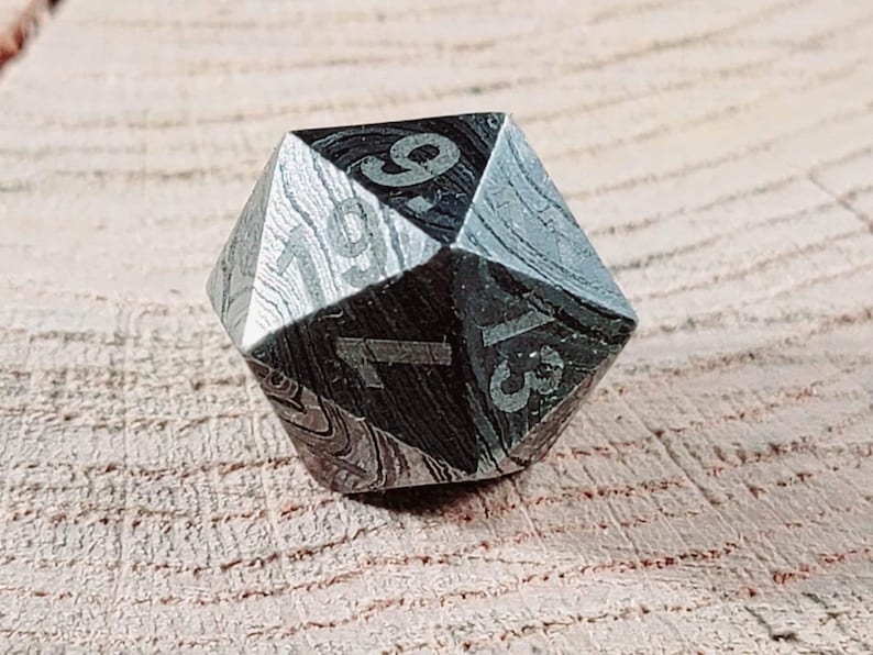 May include: A black and silver metal d20 dice with numbers 1 through 20 etched on each side. The dice has a textured, marbled pattern.