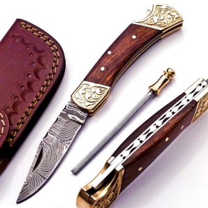 Peut inclure: Un couteau pliant de poche avec un manche en bois et une lame en acier damas. Le couteau a une mitre en laiton et un motif floral en laiton sur le manche. Le couteau est livré avec un étui en cuir.