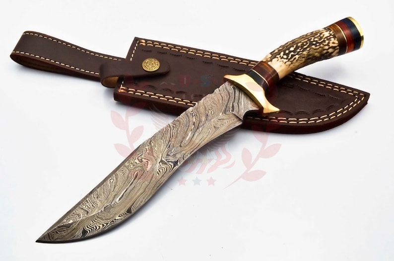 Puede incluir: Un cuchillo de caza con una funda de cuero marr&oacute;n. El cuchillo tiene una hoja de acero damasco y un mango de asta de ciervo con una virola de lat&oacute;n.