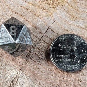 May include: A silver, eight-sided die with a patterned surface, showing the number eight, next to a US quarter.