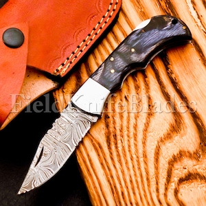 May include: A folding pocket knife with a Damascus steel blade and a dark wood handle. The knife is open and the blade is sharp. The knife is sitting on a wooden surface.