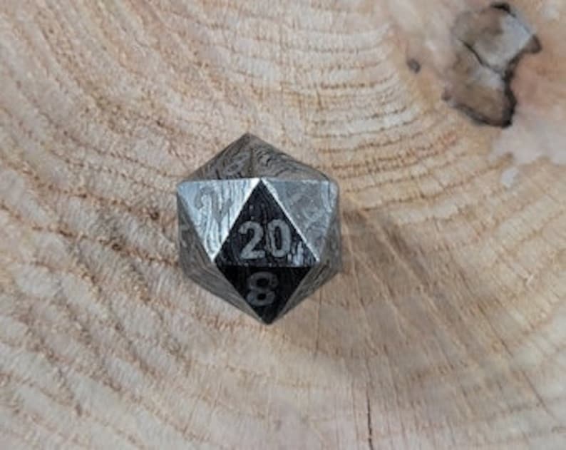 May include: A black metal 20-sided die with numbers etched on each face. The die is sitting on a wooden surface.