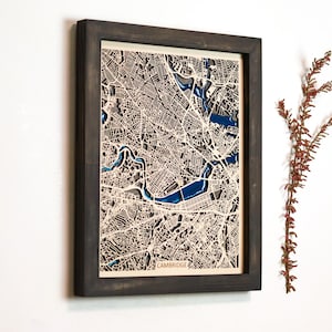 May include: Framed laser-cut map of Cambridge, Massachusetts, with a dark gray wooden frame. The map features intricate street and river details in beige and blue. The word "CAMBRIDGE" is visible at the bottom.
