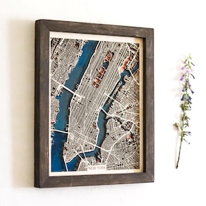May include: A framed, multi-layered wooden map of New York City featuring intricate laser-cut street grids in tan and brown tones, set against a deep blue water background. The piece is enclosed in a dark brown wooden frame with the text NEW YORK.