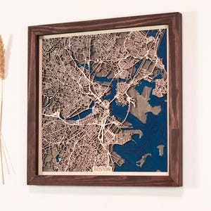 May include: A framed wooden map of Boston, Massachusetts. The map is a detailed, layered design with a dark brown frame. The city's streets and waterways are intricately carved, with a blue background representing the water. The word "BOSTON" is visible.