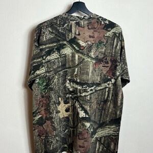 K&ouml;nnte beinhalten: Ein kurz&auml;rmeliges Camouflage-T-Shirt h&auml;ngt an einem schwarzen Kleiderb&uuml;gel. Das Muster des Shirts ist eine detaillierte Mischung aus Braun, Gr&uuml;n und Beige, die einen Wald mit Bl&auml;ttern und &Auml;sten nachahmt. Der Stoff wirkt weich und atmungsaktiv.