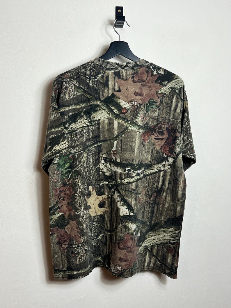 K&ouml;nnte beinhalten: Ein kurz&auml;rmeliges Camouflage-T-Shirt h&auml;ngt an einem schwarzen Kleiderb&uuml;gel. Das Muster des Shirts ist eine detaillierte Mischung aus Braun, Gr&uuml;n und Beige, die einen Wald mit Bl&auml;ttern und &Auml;sten nachahmt. Der Stoff wirkt weich und atmungsaktiv.