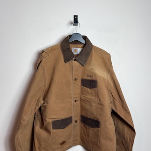 Könnte beinhalten: Braune Carhartt-Jacke mit braunem Cordkragen und dunkelbraunen Akzenten an den Taschen. Die Jacke hat vier Vordertaschen und den Namen "Senna" auf der rechten Brust gestickt. Sie hängt an einem schwarzen Kleiderbügel.
