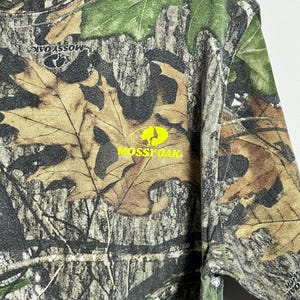May include: Camouflage long-sleeve shirt with a crew neck and the Mossy Oak logo in yellow. The shirt features a pattern of brown, green, and tan leaves and branches, with a bright yellow collar.