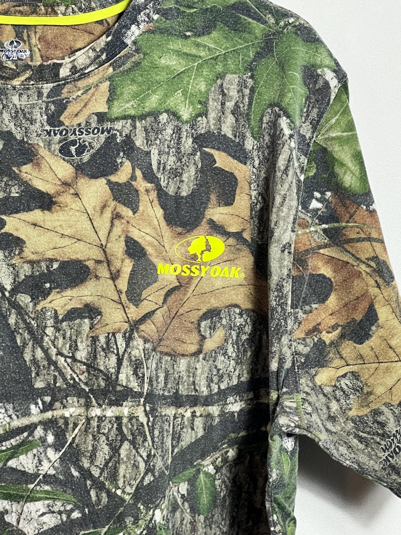 May include: Camouflage long-sleeve shirt with a crew neck and the Mossy Oak logo in yellow. The shirt features a pattern of brown, green, and tan leaves and branches, with a bright yellow collar.