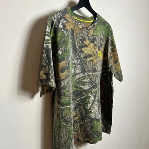 May include: A short-sleeved camouflage t-shirt hangs on a black hanger against a white wall. The shirt features a green, brown, and tan woodland camouflage pattern. A small yellow logo is visible on the chest.