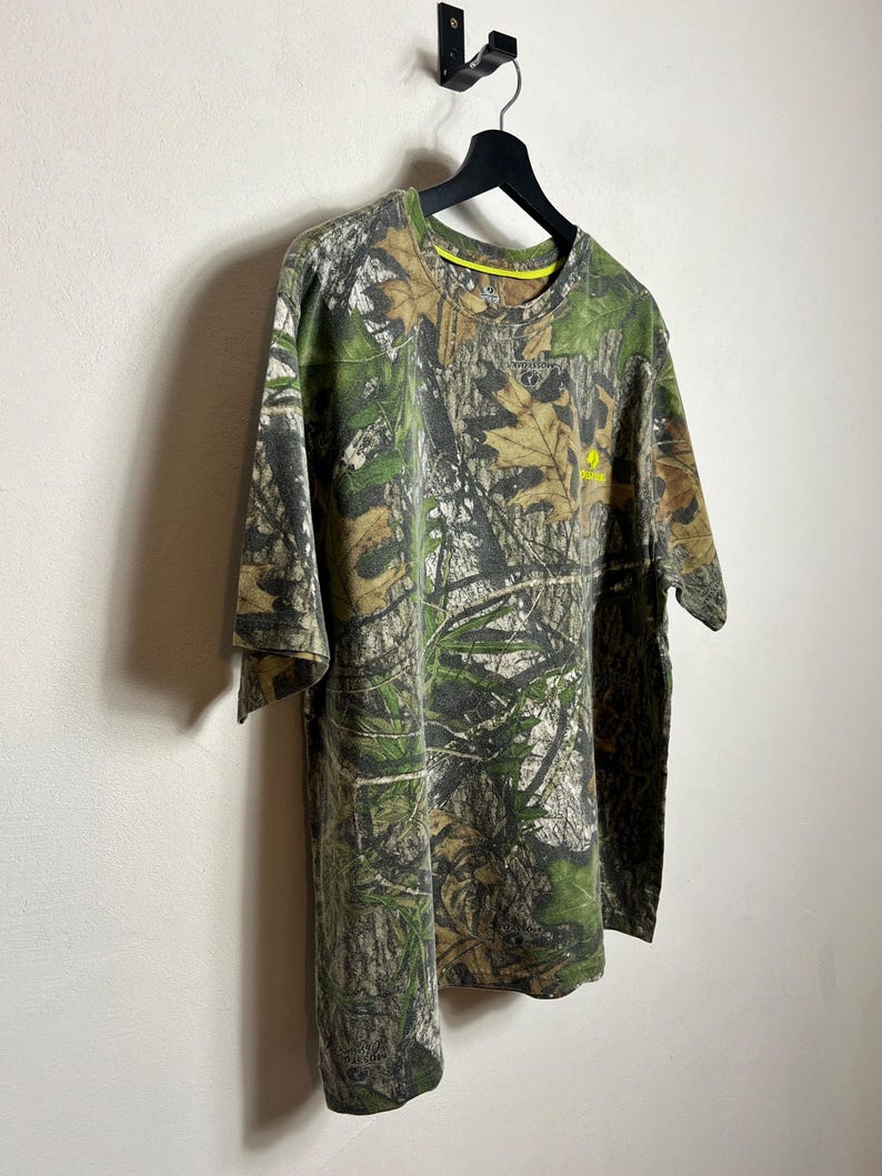 May include: A short-sleeved camouflage t-shirt hangs on a black hanger against a white wall. The shirt features a green, brown, and tan woodland camouflage pattern. A small yellow logo is visible on the chest.