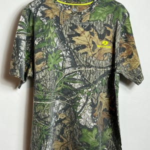 May include: A short-sleeved camouflage t-shirt with a crew neck. The shirt features a pattern of green, brown, and tan leaves and branches. The text "Mossy Oak" is printed in yellow on the front.