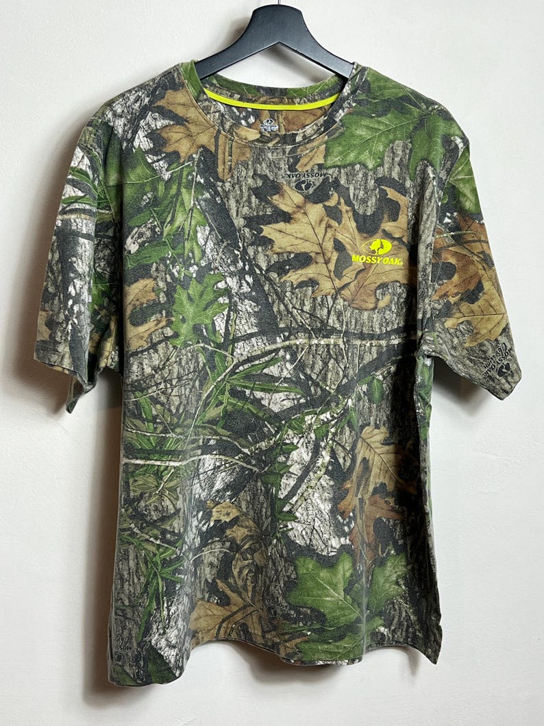 May include: A short-sleeved camouflage t-shirt with a crew neck. The shirt features a pattern of green, brown, and tan leaves and branches. The text "Mossy Oak" is printed in yellow on the front.