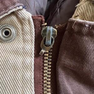 May include: Close-up of a brown and beige jacket with a gold zipper. The zipper has a metal pull with a blue finish.