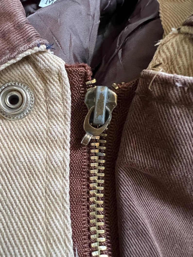 May include: Close-up of a brown and beige jacket with a gold zipper. The zipper has a metal pull with a blue finish.