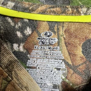 May include: Camouflage pattern t-shirt with a neon yellow neckline. The tag reads "Mossy Oak Obsession" and lists the size as XL/XG (46-48). The shirt is made of cotton and polyester.
