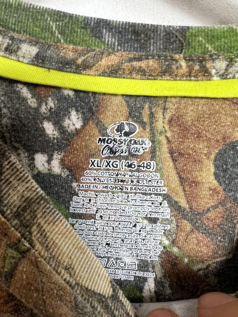May include: Camouflage pattern t-shirt with a neon yellow neckline. The tag reads "Mossy Oak Obsession" and lists the size as XL/XG (46-48). The shirt is made of cotton and polyester.