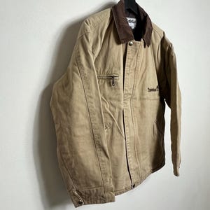May include: A tan work jacket with a brown corduroy collar and a brown embroidered logo that says "Timberland" on the left chest.