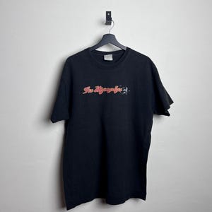 May include: Black t-shirt with the text "Fu Manchu" in a pink and red font. The shirt is hanging on a black hanger against a white wall. The t-shirt is a crew neck style.