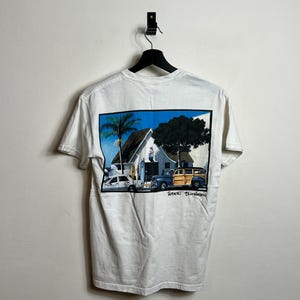 Könnte beinhalten: Weißes T-Shirt mit einem Grafikdruck einer Strandszene mit einer Palme, einem Gebäude und einem Oldtimer. Der Text "Robert Bluhm" ist unter dem Grafikdruck gedruckt.