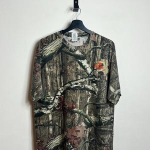 K&ouml;nnte beinhalten: Ein T-Shirt mit Camouflage-Muster und Rundhalsausschnitt, das an einem schwarzen Kleiderb&uuml;gel h&auml;ngt. Das Shirt zeigt einen detaillierten Walddruck in Braun-, Gr&uuml;n- und Beiget&ouml;nen. Ein kleines orangefarbenes Logo ist auf der rechten Seite sichtbar.