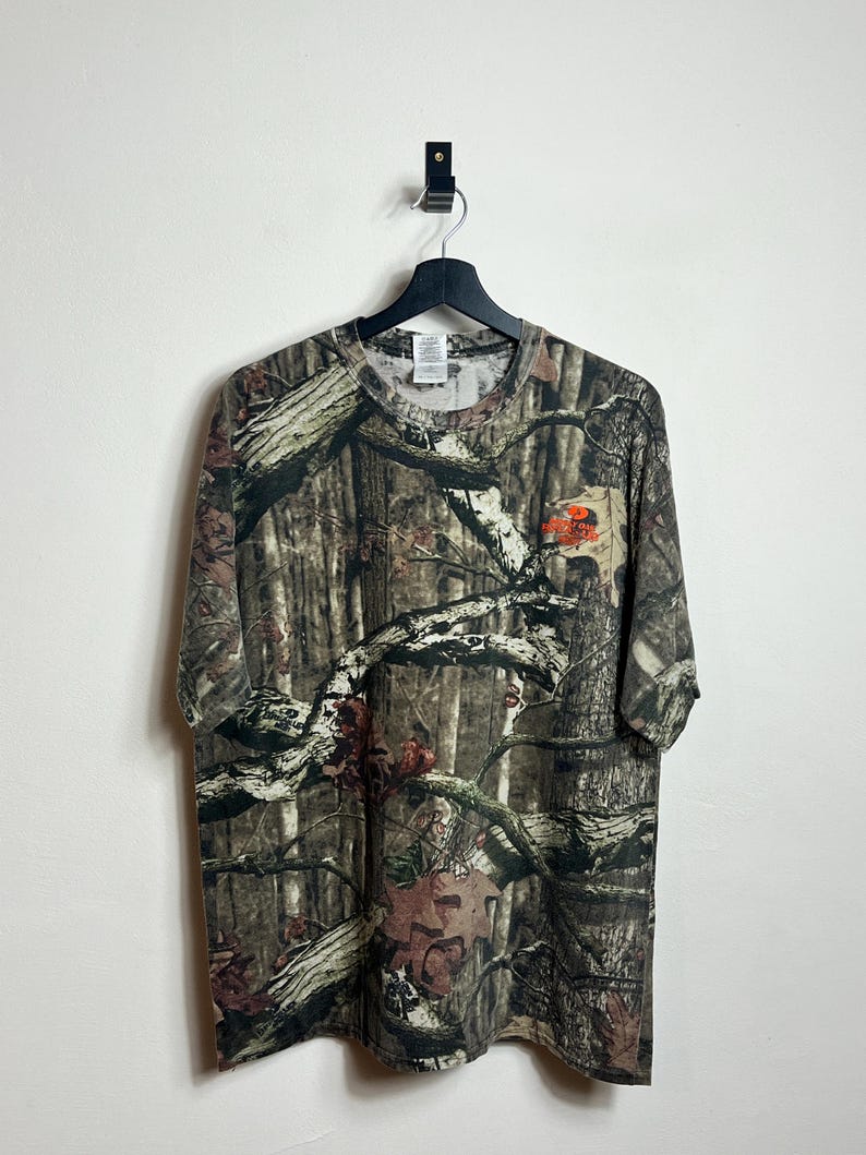 K&ouml;nnte beinhalten: Ein T-Shirt mit Camouflage-Muster und Rundhalsausschnitt, das an einem schwarzen Kleiderb&uuml;gel h&auml;ngt. Das Shirt zeigt einen detaillierten Walddruck in Braun-, Gr&uuml;n- und Beiget&ouml;nen. Ein kleines orangefarbenes Logo ist auf der rechten Seite sichtbar.