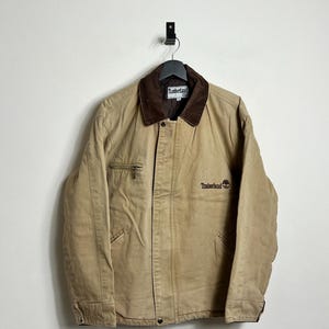 Può includere: Una giacca da lavoro beige con colletto in velluto a coste marrone e un logo ricamato marrone che dice "Timberland" sul lato destro del petto.