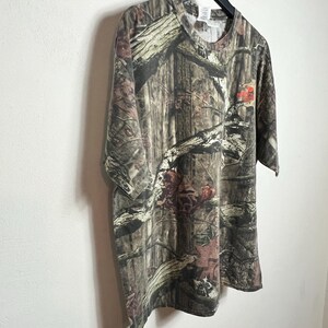 K&ouml;nnte beinhalten: Ein kurz&auml;rmeliges Camouflage-T-Shirt mit Rundhalsausschnitt. Das Shirt weist ein Muster aus braunen, gr&uuml;nen und beigen Farbt&ouml;nen auf, das eine Waldumgebung nachahmt. Das Shirt h&auml;ngt an einem schwarzen Kleiderb&uuml;gel vor einer wei&szlig;en Wand.