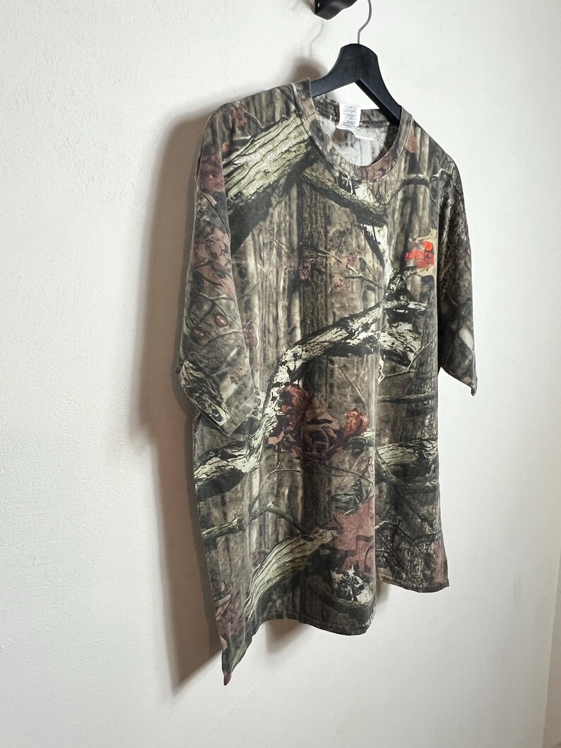 K&ouml;nnte beinhalten: Ein kurz&auml;rmeliges Camouflage-T-Shirt mit Rundhalsausschnitt. Das Shirt weist ein Muster aus braunen, gr&uuml;nen und beigen Farbt&ouml;nen auf, das eine Waldumgebung nachahmt. Das Shirt h&auml;ngt an einem schwarzen Kleiderb&uuml;gel vor einer wei&szlig;en Wand.