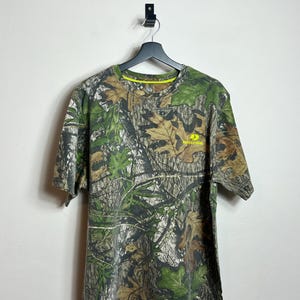 May include: A short-sleeved camouflage t-shirt with a crew neck. The shirt features a pattern of green, brown, and tan leaves and branches. The brand name "Mossy Oak" is printed in yellow on the right chest.