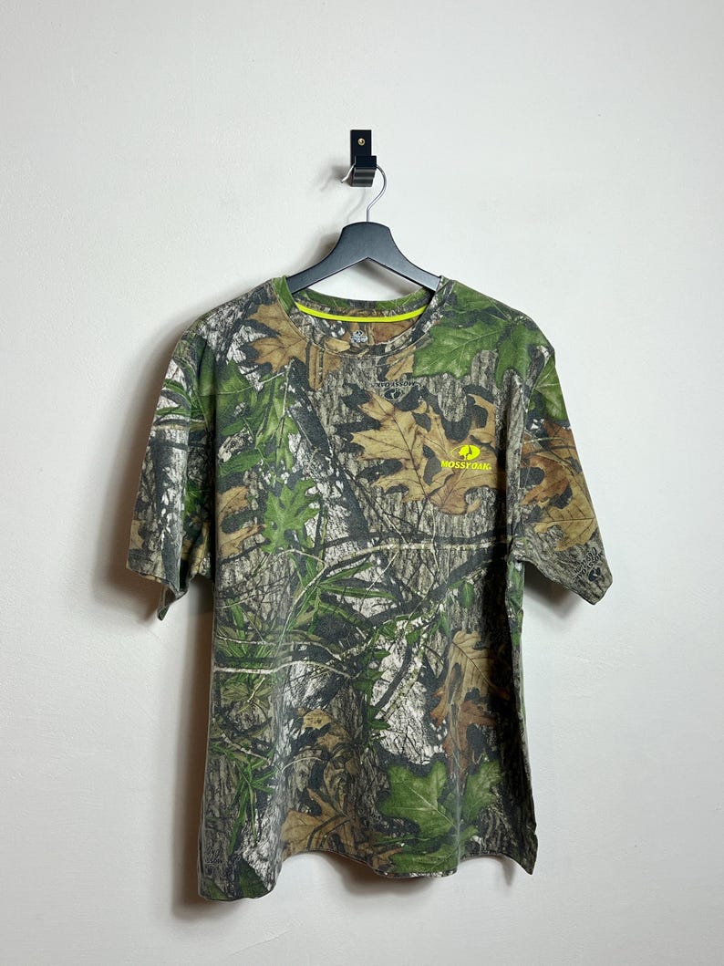 May include: A short-sleeved camouflage t-shirt with a crew neck. The shirt features a pattern of green, brown, and tan leaves and branches. The brand name "Mossy Oak" is printed in yellow on the right chest.