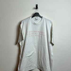 Peut inclure: T-shirt blanc avec un imprimé graphique rose qui dit "J'ai le secret d'être toutes choses à tous les peuples."