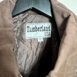 May include: A brown corduroy jacket with a brown corduroy collar and a brown quilted lining. The jacket has a label that says "Timberland USA" and "M".