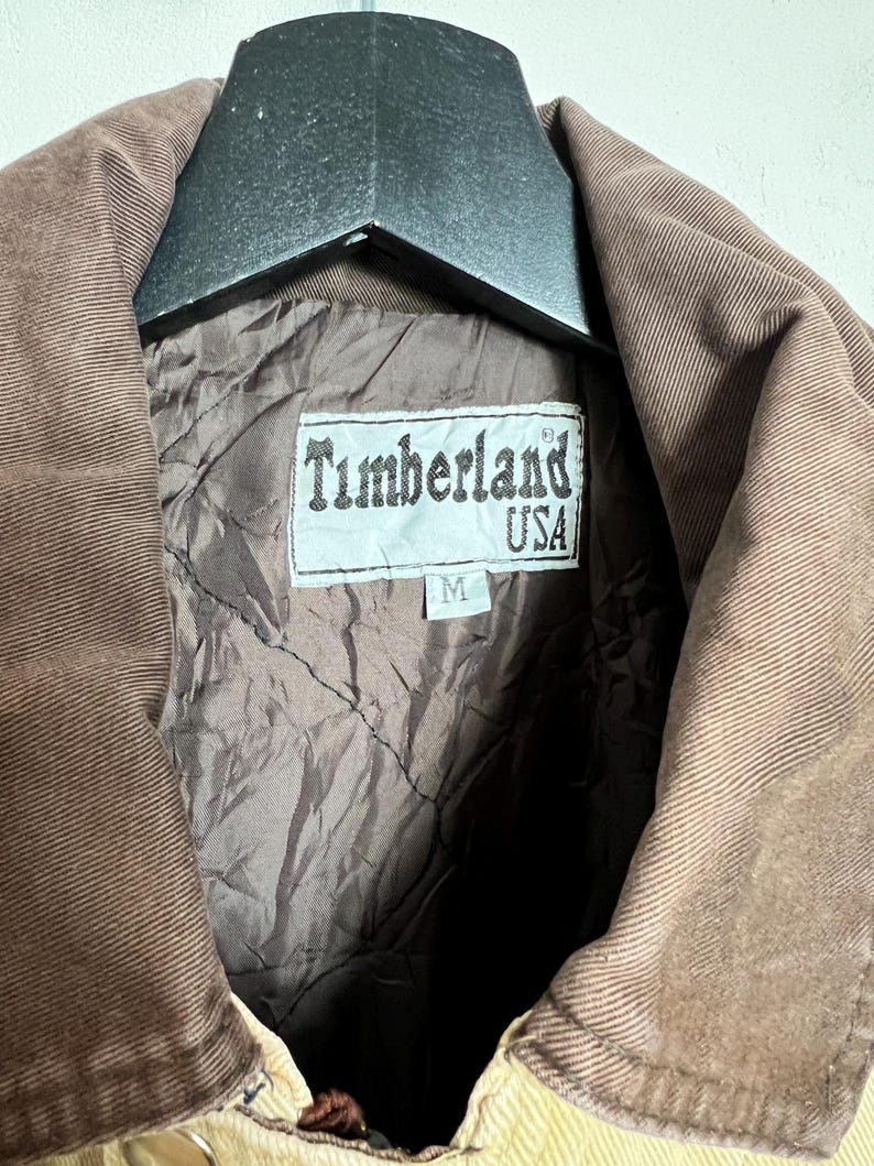 May include: A brown corduroy jacket with a brown corduroy collar and a brown quilted lining. The jacket has a label that says "Timberland USA" and "M".