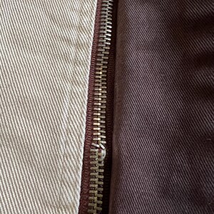 May include: Close-up of a zipper on a garment. The zipper is silver and has a brown fabric backing. The fabric on the left side of the zipper is a light beige color, while the fabric on the right side is a dark brown color.